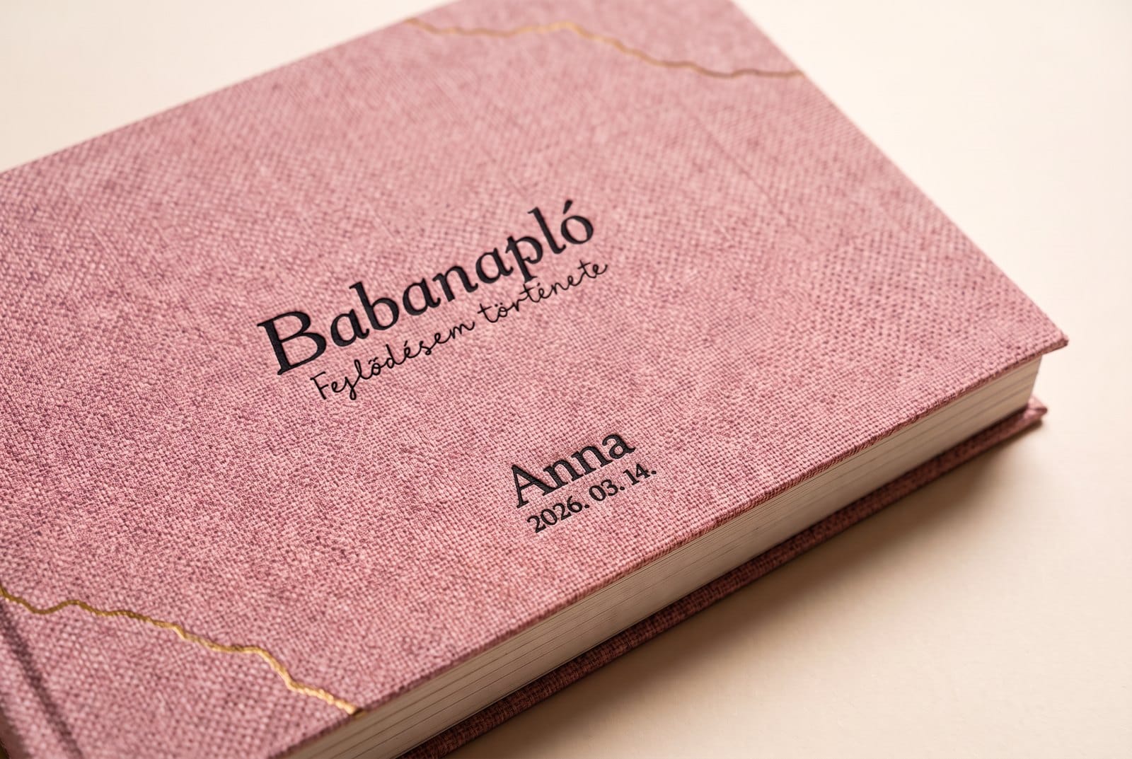 Púder rózsaszín lenvászon Babanapló borító, a cím alatt kicsi, fekete gravírozás: Anna és 2026. 03. 14.